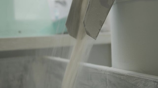 Flour is being poured from a metallic chute into a white container in a bakery. The scene shows the process of grinding flour.