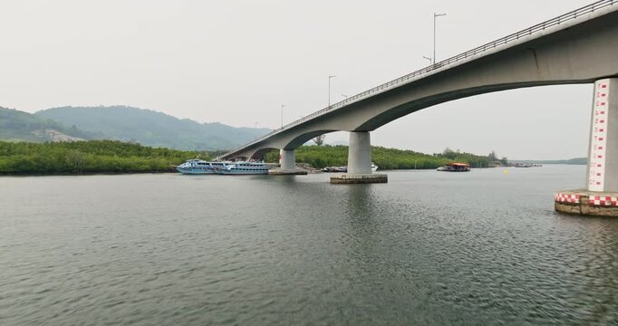 Siri Lanta Bridge connects Koh Lanta Noi to mainland, passenger ferry boats docked along calm water and lush green mangroves lining the riverbanks under a cloudy sky. Aerial view drone flight zoom out
