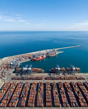 Aerial view of a busy container port terminal with stacked cargo boxes, heavy cranes, and large cargo ships docked along the blue sea under a clear sky.