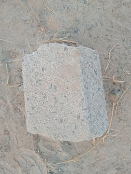 Concrete paving block on dusty ground. Single gray interlocking stone lying on dry dirt with sparse straw. High angle view of construction material left on sandy soil outdoors.