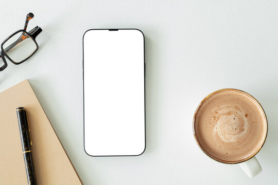 Top view flat lay of modern smartphone mockup with blank white screen on clean desk alongside coffee cup, notebook, pen, and eyeglasses. Minimalist workspace template for design.