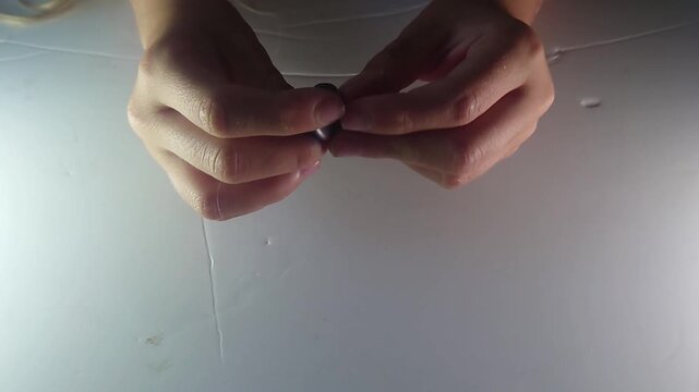 An overhead close-up shot captures hands carefully threading a piece of cord or string through the center hole of a dark clay spinning disc toy on a white background during a craft demonstration.