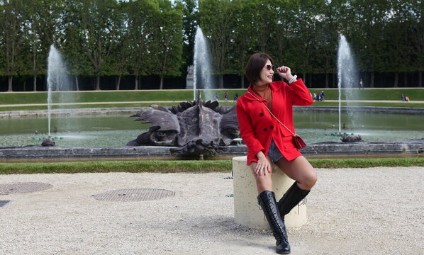 An Asian woman wearing a red jacket, brown turtleneck, and knee-high boots sits on a stone post smiling near a large ornamental pond