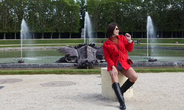 An Asian woman wearing a red jacket, brown turtleneck, and knee-high boots sits on a stone post smiling near a large park fountain basin