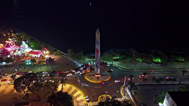 Nighttime high-angle aerial drone shot orbiting Obelisco Macho monument, street traffic roundabout, and illuminated Juan Baron amusement park at Santo Domingo Malecon.