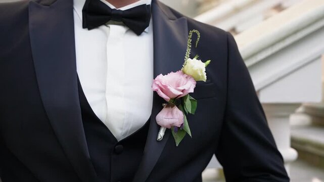 Cinematic close-up shot of a groom in a classic black tuxedo with a pastel boutonniere, standing before ornate staircase. Sharp focus, natural light, wedding elegance, intimate detail.
