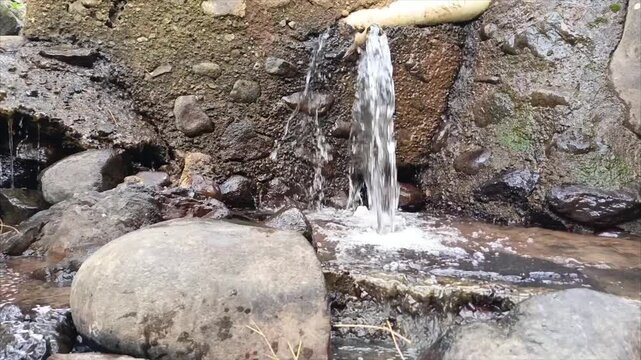 Clear natural water flowing from bamboo pipe into rocky stream with stones in tea plantation. Small waterfall from bamboo aqueduct in countryside. Natural water source and irrigation concept for backg