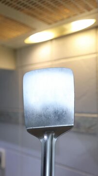 Close-up of a metal spatula with blue handle used for flipping chapatis in a bright kitchen with overhead lighting. Warm, domestic cooking atmosphere.