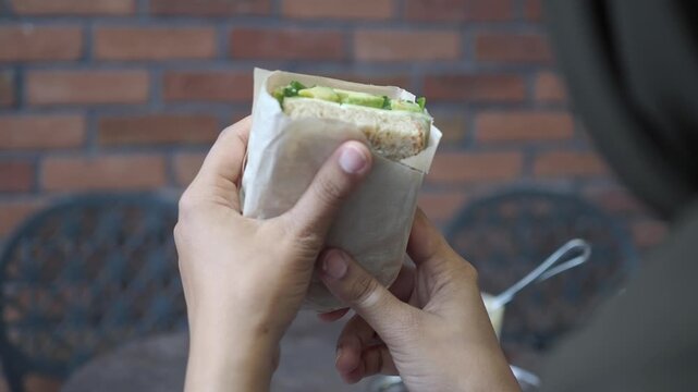 A young woman holds a sandwich wrapped in paper. It is at a busy cafe
