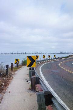  A winding waterfront road features sharp chevron traffic signs alongside a pedestrian sidewalk overlooking a calm Mission Bay under cloudy gray morning skies, San Diego, CA