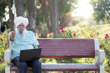 Fototapeta premium Indian senior man showing thumbs up while working on laptop at park