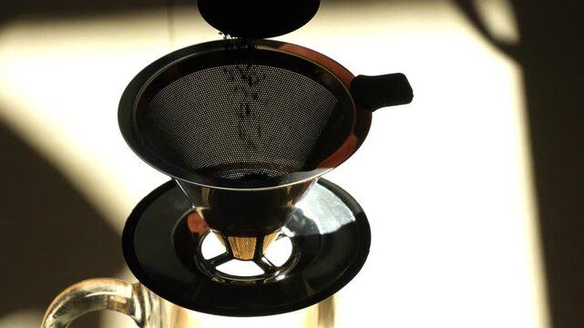 Pouring freshly ground coffee powder into a stainless steel pour-over cone dripper filter, backlit high contrast close-up shot.
