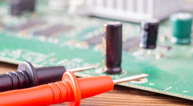 Red and black multimeter probes lying in front of a blurred green electronic circuit board