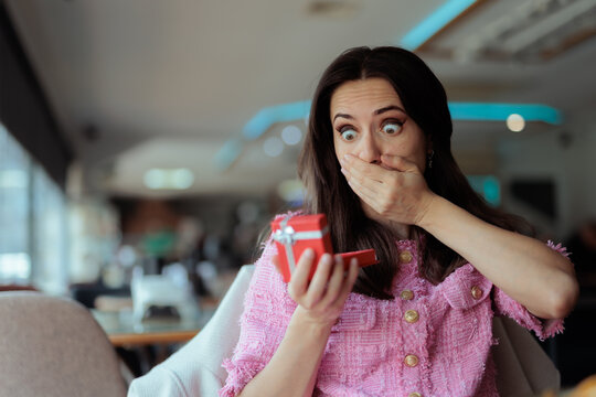 Surprised Young Woman Opening Red Gift Box Inside Modern Restaurant. Astonished brunette female gasps while holding small jewelry present box indoor
