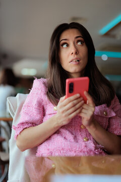 Wondering Woman Holding Smartphone While Looking Away. Beautiful young brunette woman in stylish pink jacket holding red phone with a thoughtful facial expression