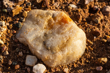 Smooth white vein quartz boulder resting on raw gravel ground, sunlit natural mineral texture with golden highlights in a mining quarry landscape surface close-up perspective © PhotoChur