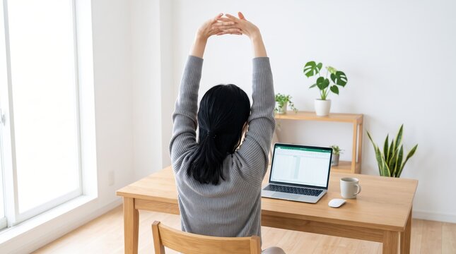 Rear view of young woman sitting at wooden desk stretching arms upwards to relieve stress and fatigue while working from home, demonstrating healthy workplace habits