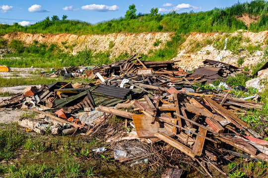 Heap of discarded construction waste and rotting lumber piled up at an abandoned quarry site amidst lush green hills under a bright blue sky, environmental pollution concept