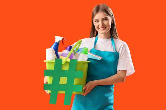 Female janitor holding bucket with cleaning supplies and hashtag sign on color background