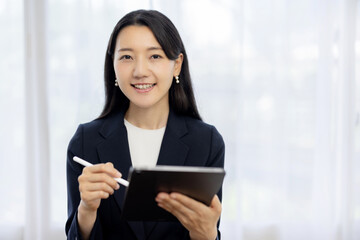 Obraz premium Young Japanese businesswoman in a suit using a tablet and smiling at the camera at a desk by the window