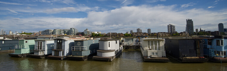 Obraz premium Colorful residential houseboats moored along the River Thames in London, England, with modern high-rise buildings and the city skyline visible in the background.