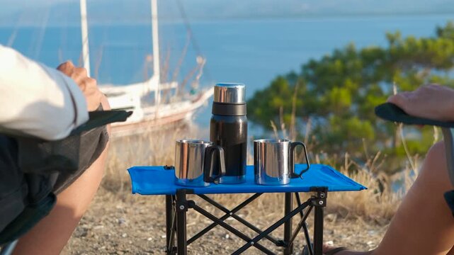 Tourists enjoy morning coffee by the lake. The couple carefully places a thermos and metal cups on a small, portable camping table. Relaxation concept of morning coffee at camping by the sea.
