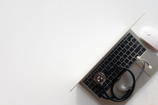 Top view of a black medical stethoscope on a silver laptop keyboard and white mouse on a white desk with large copy space. Concept of telemedicine, health tech, and online doctor consultation.