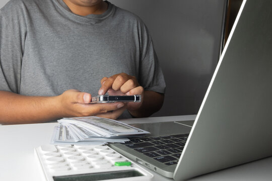 Man using a smartphone with a stack of US dollar prop money, calculator, and laptop on desk. Concept of making money online, mobile banking, freelance income, and financial investment.