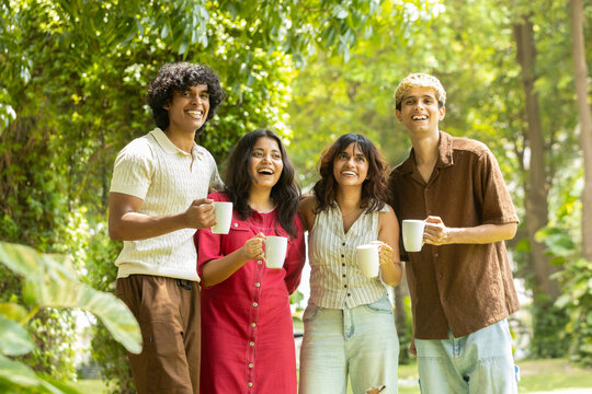 Group of diverse indian asian Gen z young adult friends holding coffee mugs laughing and embracing each other outdoors, sharing happy moment and, friendship in summer park. Togetherness. Youth culture