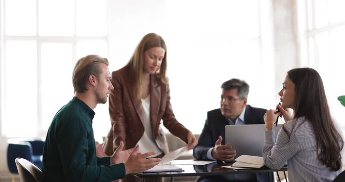 Female business trainer guiding group conversation giving paper materials to speaking team member supporting trainee answering question during professional development workshop in corporate office