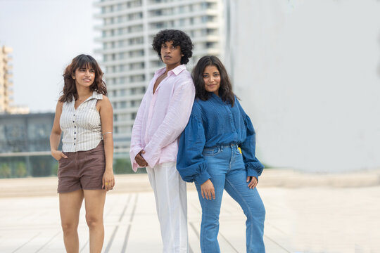 Three young diverse indian asian Gen Z adults standing confidently posing wearing casual cloths on an urban promenade, with modern residential buildings backdrop. Fashion and lifestyle.