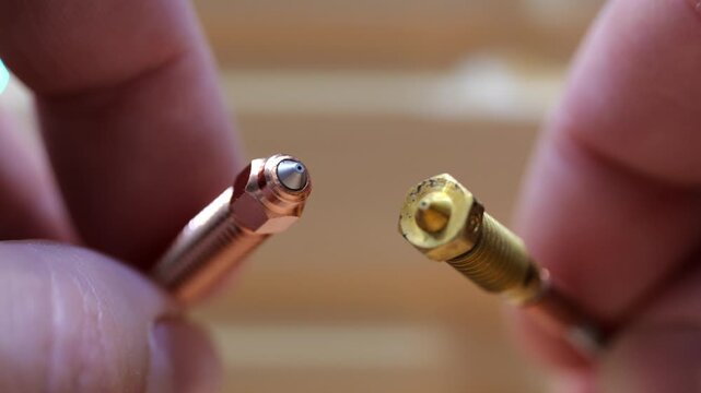 Close-up of male fingers holding and comparing two different 3D printer nozzles made of hardened steel and brass