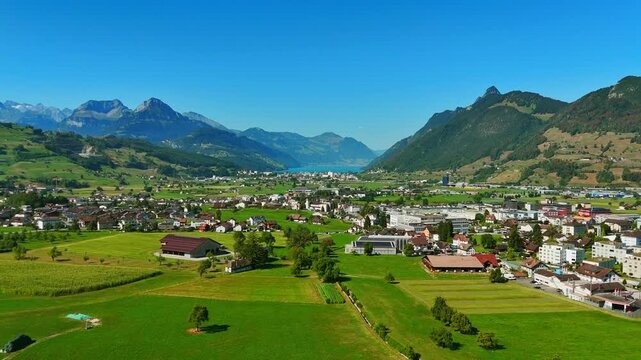 Aerial view of Schwyz, Switzerland, with green farmland, compact neighborhoods and dramatic Alpine mountains framing the valley, with a distant lake under clear summer skies.