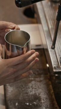 Vertical video of barista pouring milk into stainless steel pitcher and frothing it with espresso steam wand. Suitable for cafe marketing, beverage commercials, and menu promo