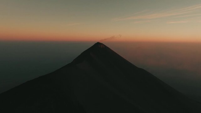 Aerial drone view of Fuego erupting at sunrise, dramatic ash cloud and colorful dawn. Intense orange hues color the sky as volcanic smoke drifts above sharp, dramatic slopes.