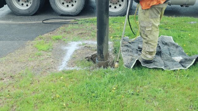 A worker uses a hydro vac wand to expose a marked section of ground beside a road, clearing soil around a vertical pipe for directional drilling.