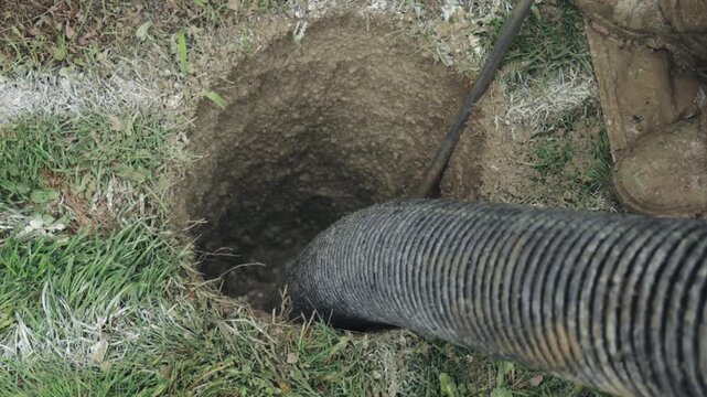 A hydrovac hose inserted into an open pit on a grassy verge while a worker clears soil to expose and install electrical conduit for street lighting; a boot and hand tool are visible at the hole's edge