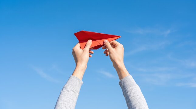 Male hands holding red paper airplane against clear blue sky, representing freedom, creativity, childhood dreams, startup launch, innovation, travel aspirations, and launching new