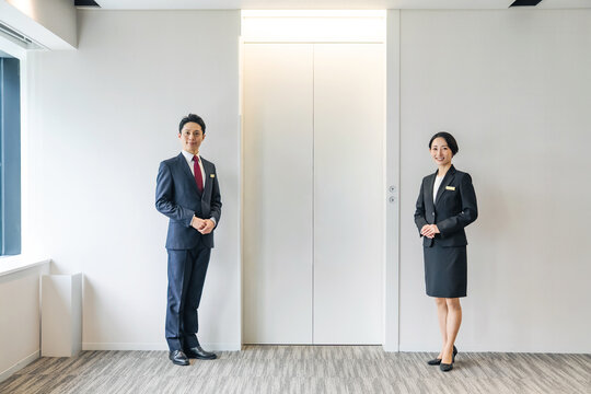 エレベーターの前に立つ男女。受付、出迎え、案内、スタッフ、誘導、ホール、ホテル、ホテリエ、点検、保守