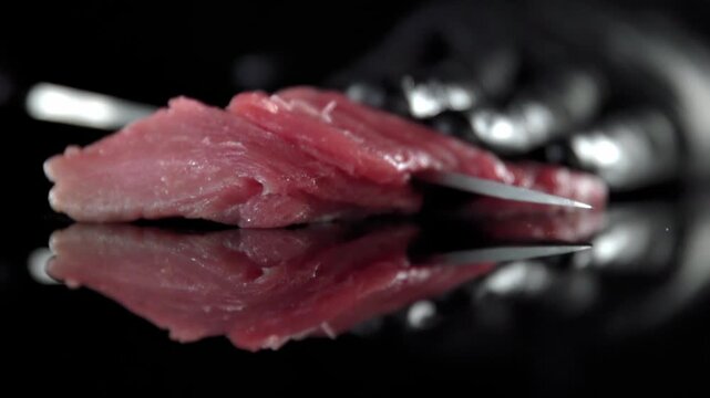 Chef hand in black glove slicing fresh raw red meat with professional steel knife on dark reflective surface.