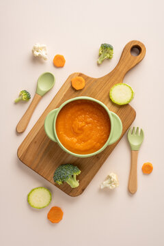 Baby's first feeding with healthy carrot puree in a baby bowl on a wooden cutting board on vertical beige backdrop