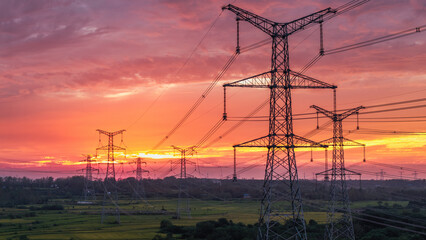 Silhouetted high voltage transmission towers stretch across rural fields under a vibrant sunset sky, part of power infrastructure. © tai