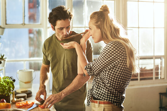 Couple, spoon and feed in kitchen with meal prep, help partner and taste test for bonding together. People, dating and cooking food in home with lunch recipe, relationship connection and wellness.