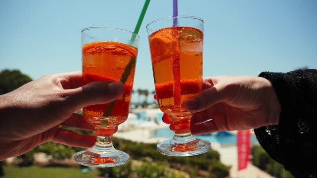Two glasses of orange spritz with ice and citrus slices held outdoors on a sunny summer day. Refreshing cocktail scene with a scenic resort background. High quality 4k footage