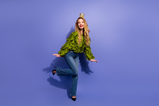 Charming young female model poses in green ruffled blouse and jeans with crown against purple background