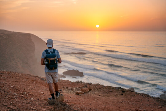 Man with backpack watching sunset over ocean at Legzira in Morocco
