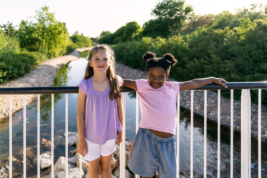 Two cheerful young girls, one caucasian and one african american, standing on a bridge, enjoying summer day outdoors