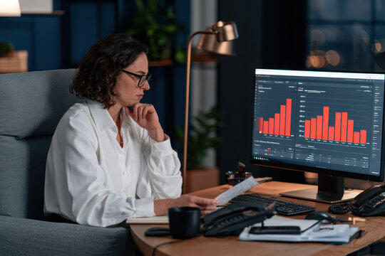 Female business analyst in white blouse reviews financial reports while seated at a desk with a computer displaying bar graphs in a modern office setting