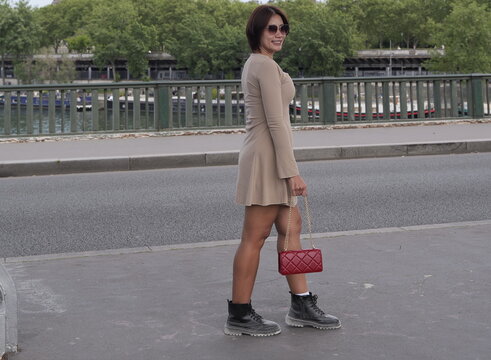An Asian woman wearing a beige long-sleeved mini dress, sunglasses, and dark boots holds a red chain-strap purse while walking along a bridge pavement