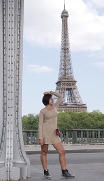 An Asian woman in a beige mini dress and boots rests her hand on her head while standing near a metal pillar bridge with the Eiffel Tower rising in the background